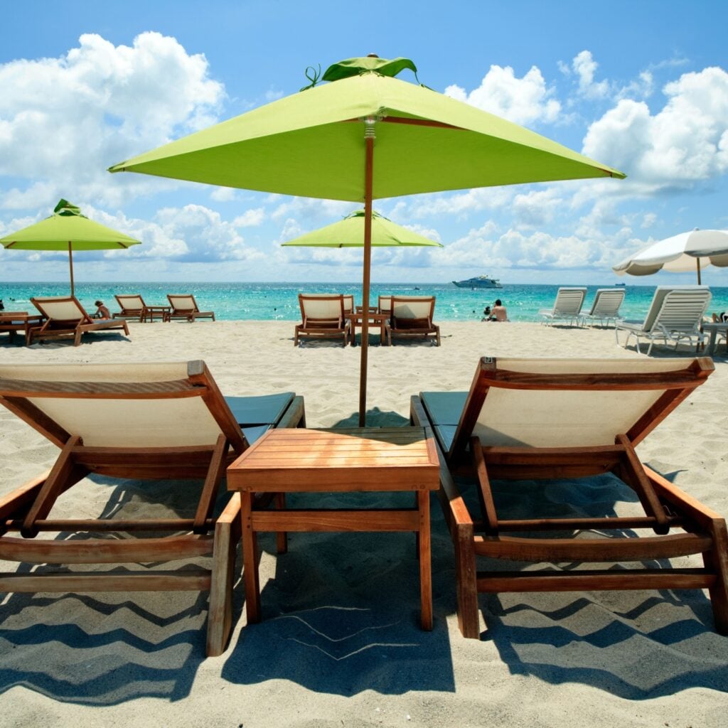 Wooden lounge chairs and green umbrellas line the sunny white-sand with clear turquoise water, capturing a perfect tropical day by the ocean in South Beach Florida.