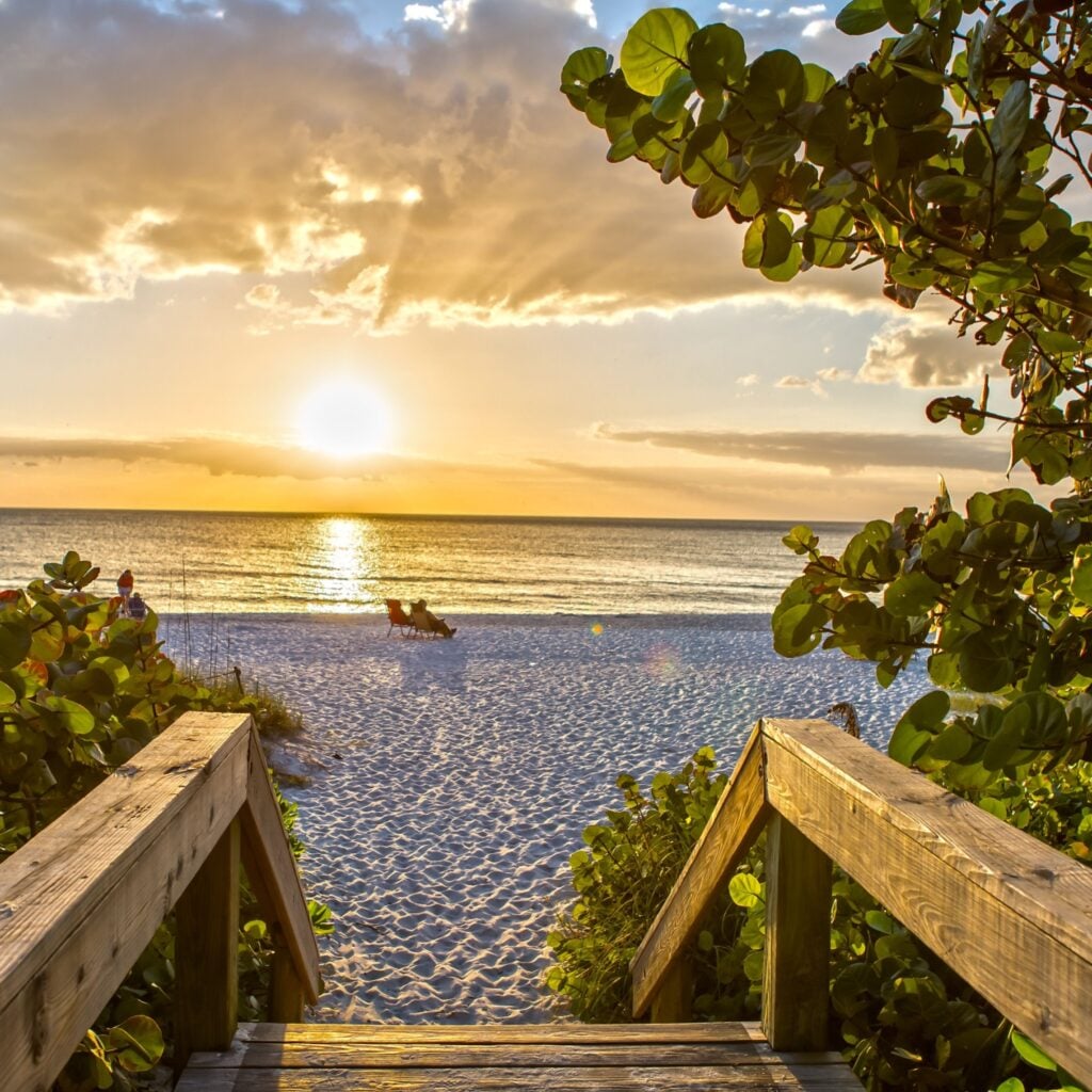 Wooden steps lead to soft white sand and calm Gulf waters at Naples Beach, with a golden sunset casting a warm glow over this peaceful coastal scene.