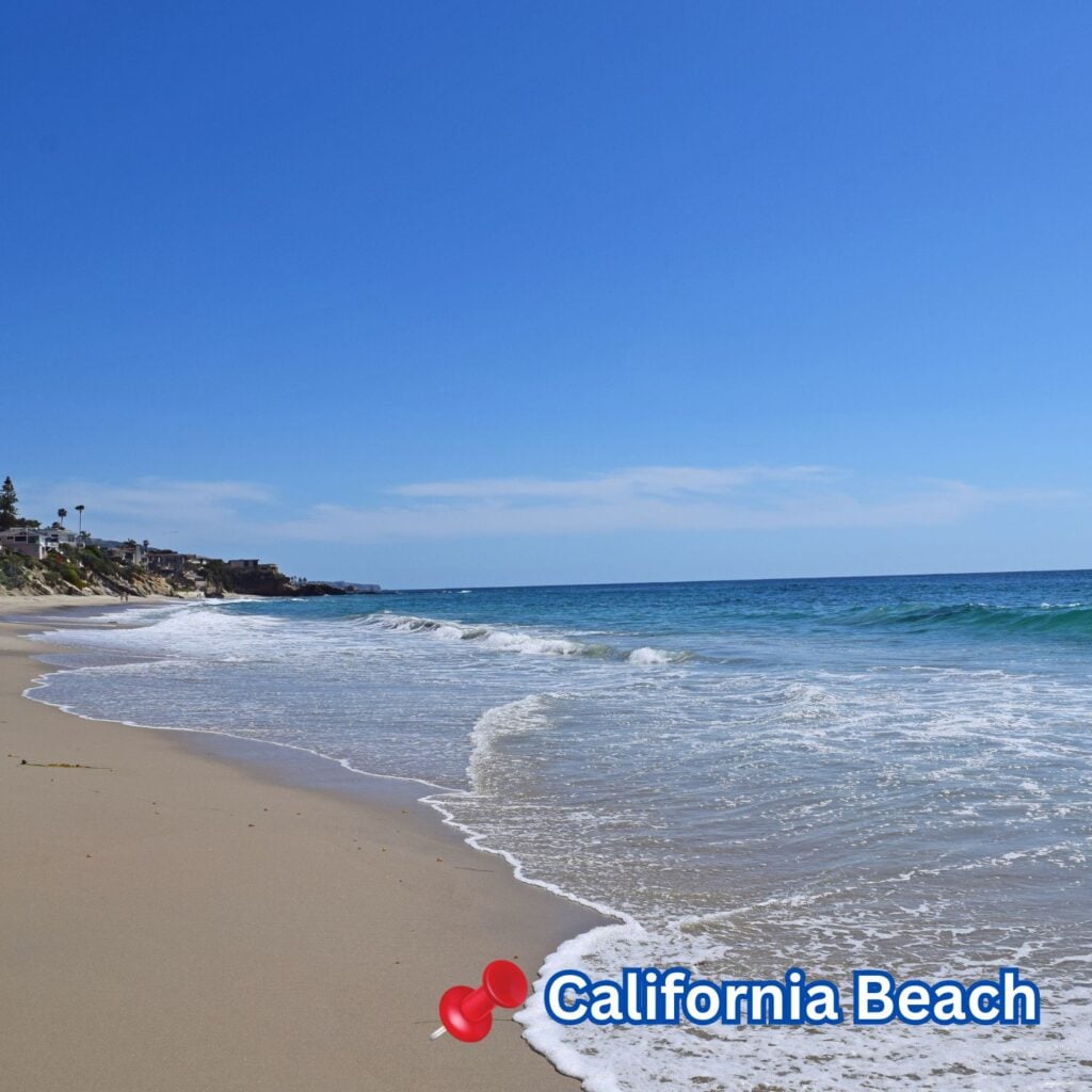 Waves gently roll onto a sandy California beach under a clear blue sky, with coastal homes and palm trees lining the distant shoreline.