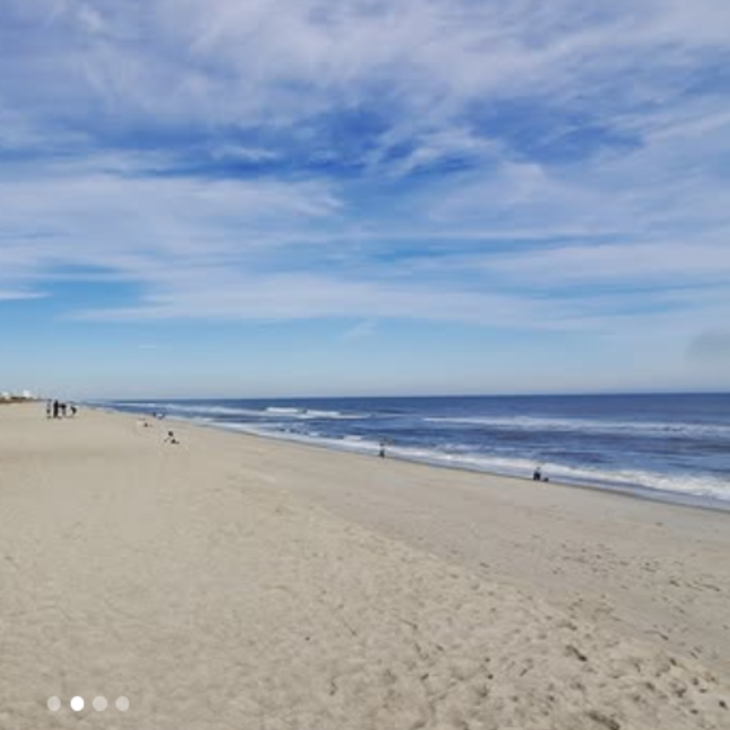 Wide sandy beach with gentle waves rolling onto Kure beach shore under a partly cloudy blue sky. Small groups of people walk and relax along the peaceful shoreline.
