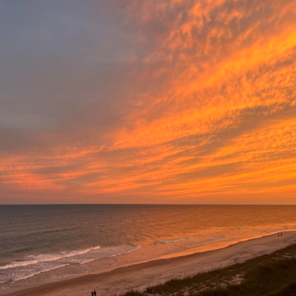 A glowing orange sunset lights up the sky over Pine Knoll Shores, with gentle waves reaching the sand and a few people walking along the peaceful beach.
