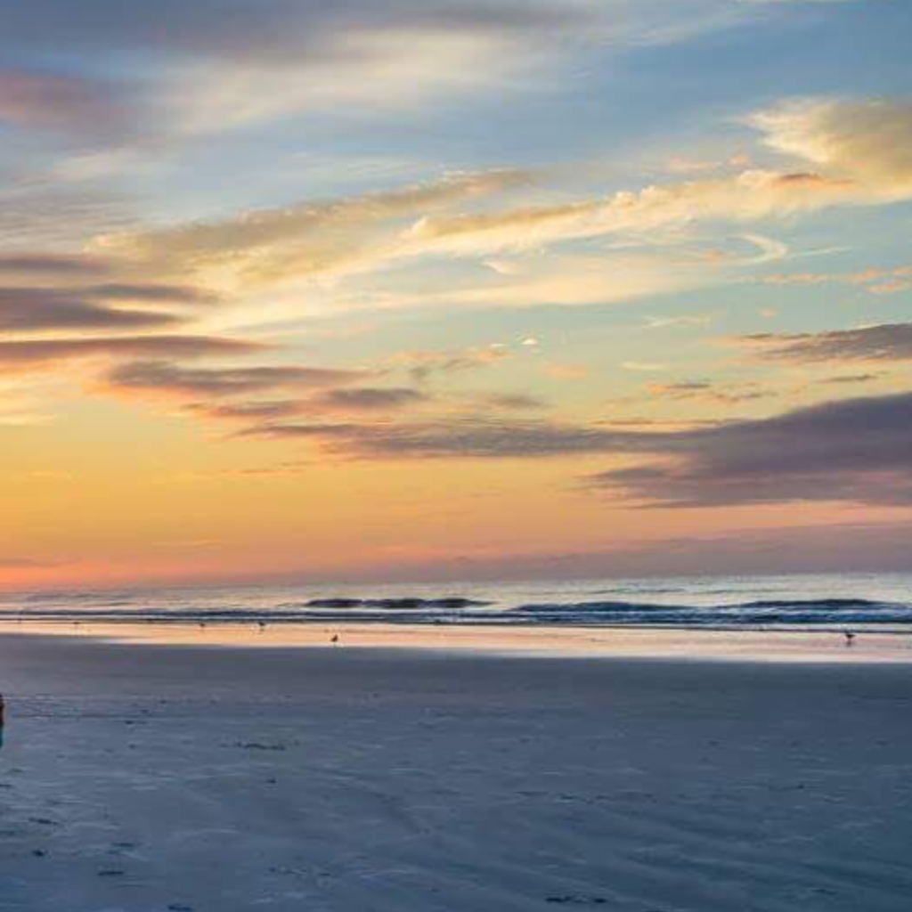 A calm beach scene at sunset with soft waves, a wide sandy shoreline, and a sky painted in warm orange, pink, and purple tones fading into twilight.