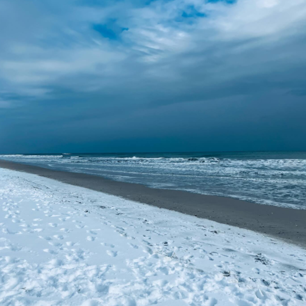 Waves roll onto surf city beach under a moody blue sky. The sand appears snow-covered with scattered footprints along the shoreline.