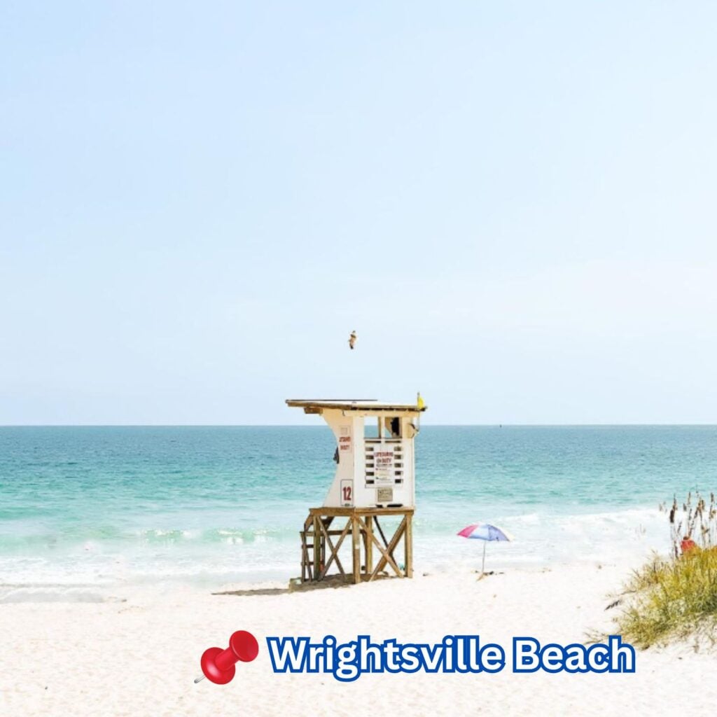 A lifeguard tower stands on the white sand of Wrightsville Beach, with calm turquoise waves, a bright sky, and a lone umbrella nearby completing the scene.