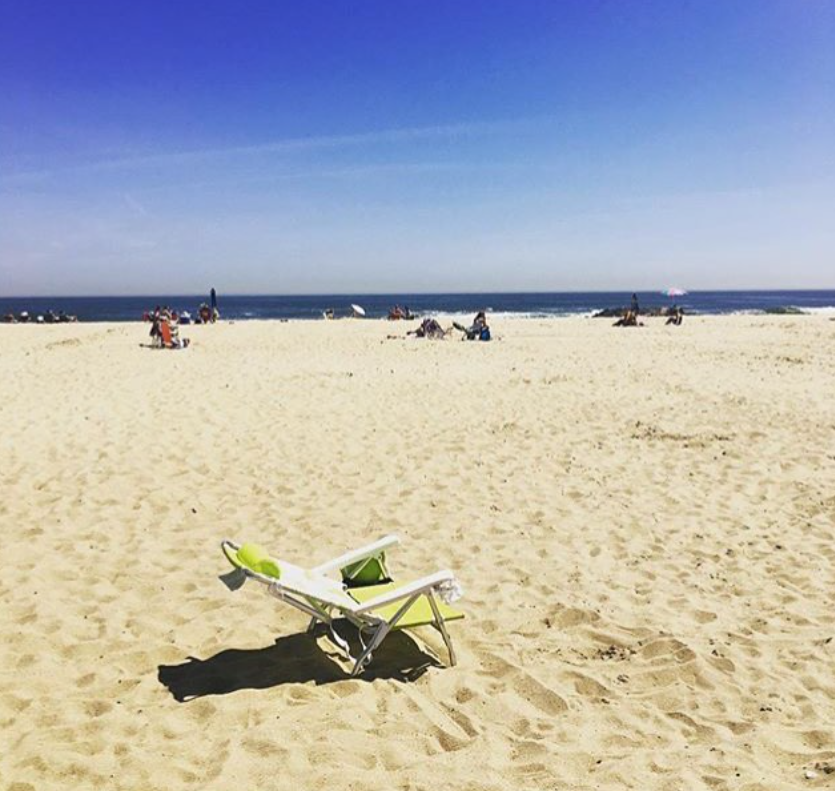 Sunny belmar beach scene with a lone green beach chair on golden sand, people relaxing in the distance near the calm blue ocean under a clear sky.









