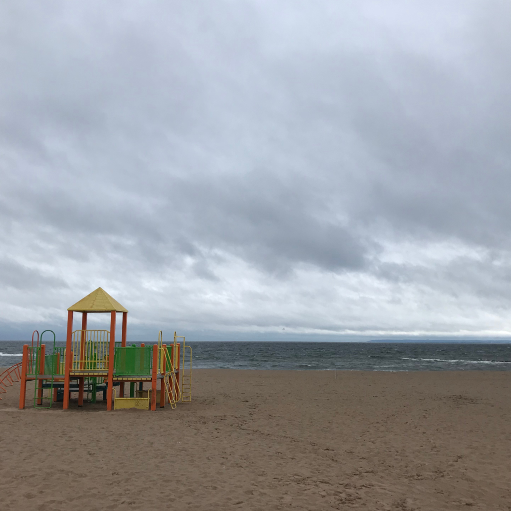 A colorful playground sits on the sandy shore at Coney Island Beach in Brooklyn under a cloudy gray sky, with waves rolling in from a choppy ocean.