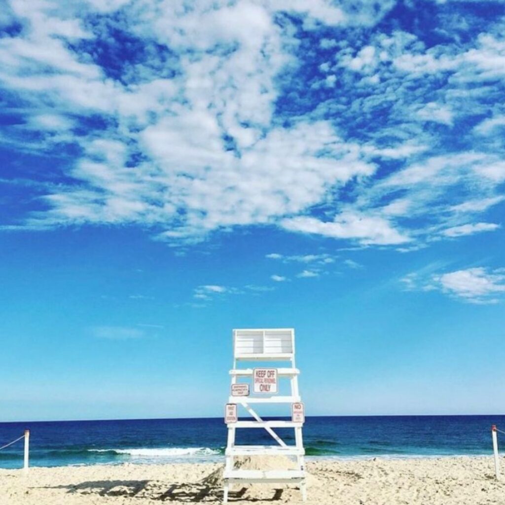 A white lifeguard chair stands on the sandy shore under a vibrant blue sky with scattered clouds, facing the calm, deep blue ocean waves.