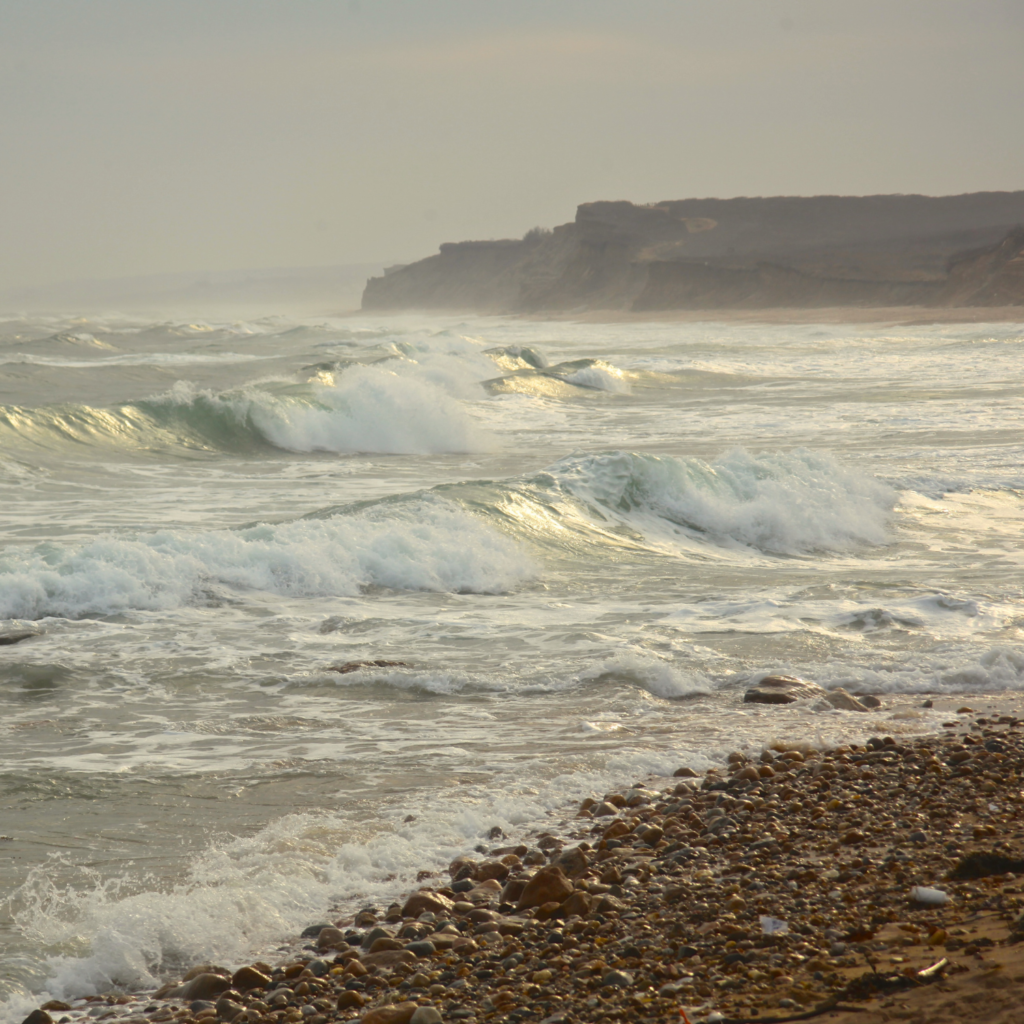 Waves crash along the rocky shoreline at Ditch Plains Beach in Montauk, with misty cliffs in the distance and a cloudy sky creating a moody coastal scene.