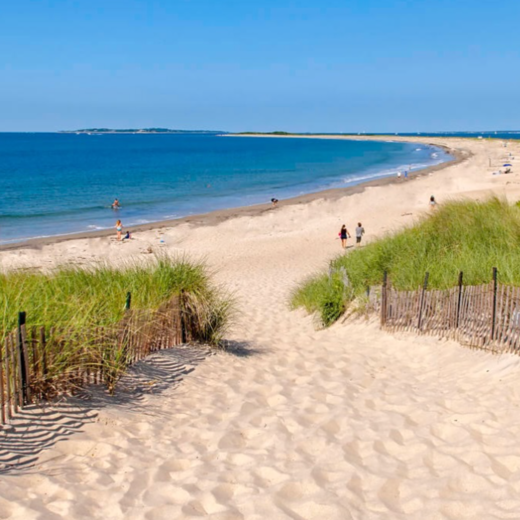A sandy trail framed by beach grass and wooden fencing leads to East Beach in Watch Hill, RI, where gentle waves meet the curved shoreline under a clear blue sky.