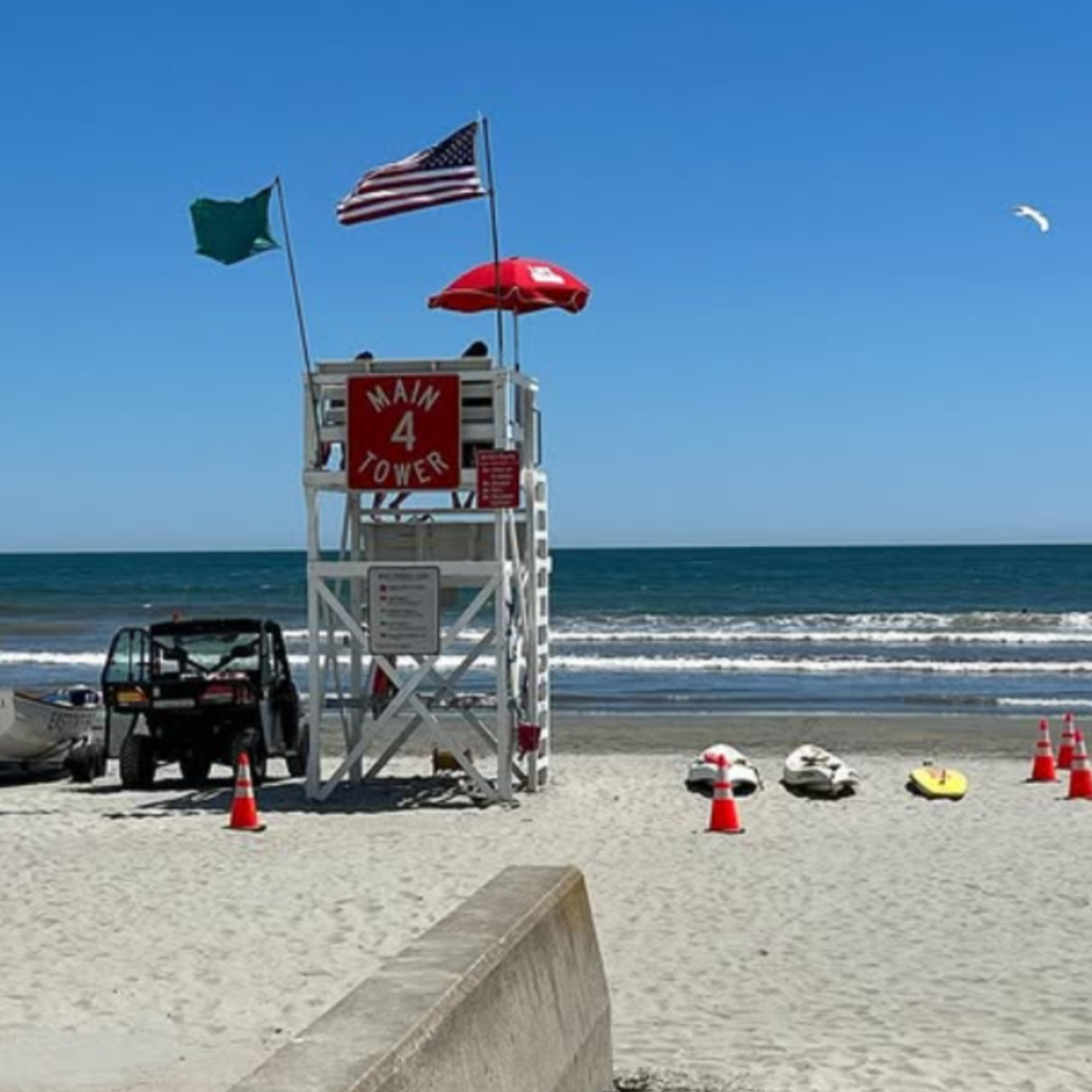 Lifeguard tower with American and green flags stands on white sand at Easton’s Beach in Newport, RI, with rescue boards, cones, and calm ocean waves nearby.