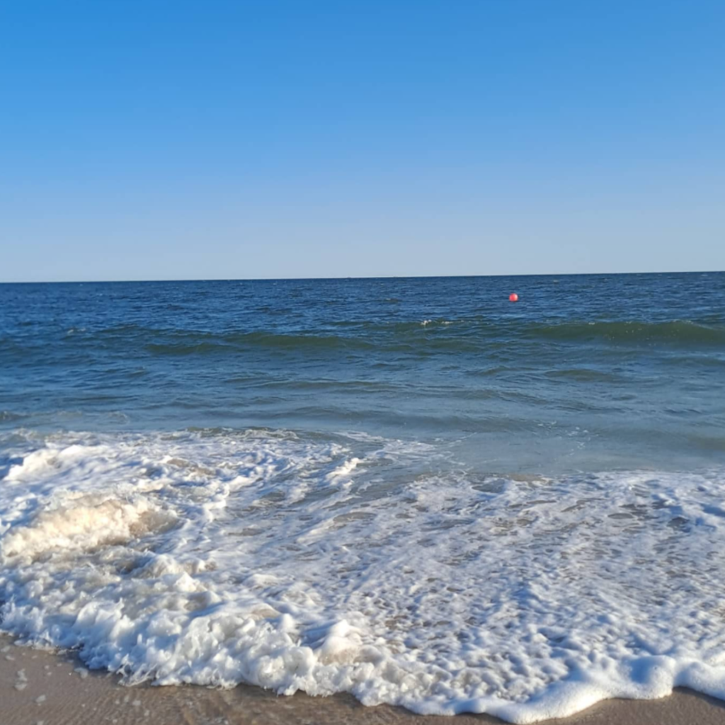 Waves gently crash on the shore at Flying Point Beach in Southampton, with deep blue water, soft foam, and a red buoy floating in the distance.