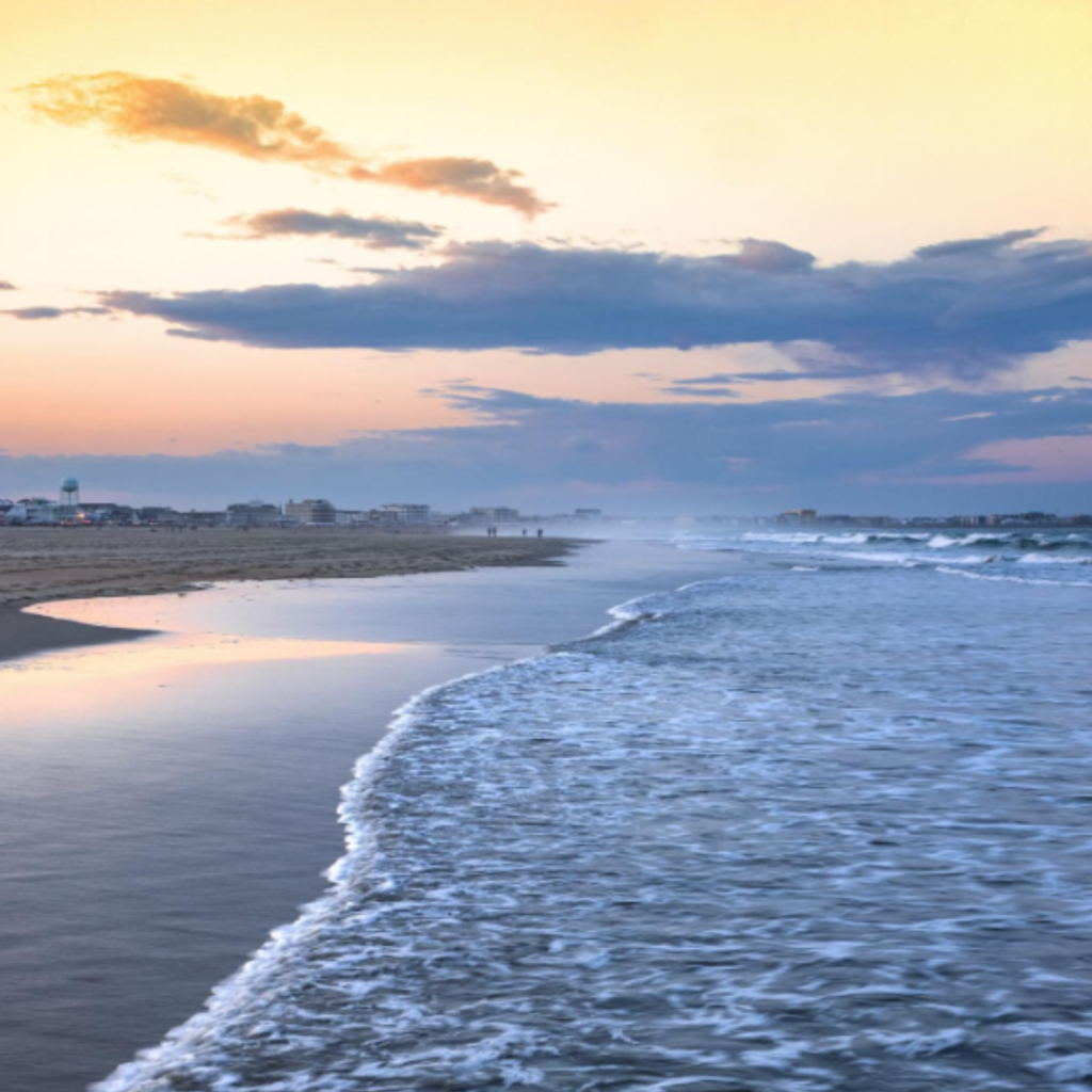 Soft waves roll onto the shore at Hampton Beach in Hampton, NH, during sunset, with pastel skies, reflections on wet sand, and beachfront buildings in the distance.