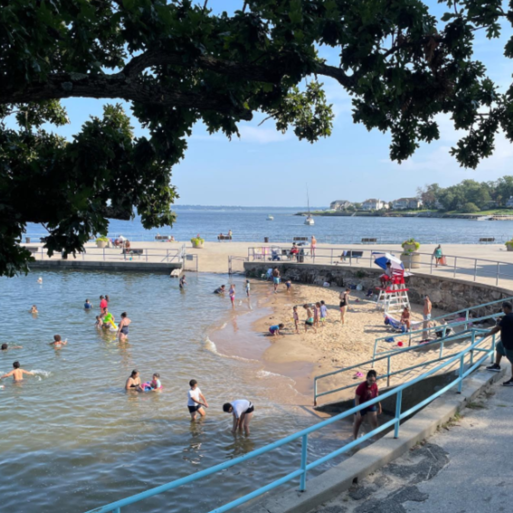 Families enjoy the calm water and small sandy area at Hudson Park and Beach in New Rochelle, with a lifeguard on duty and sailboats in the background.