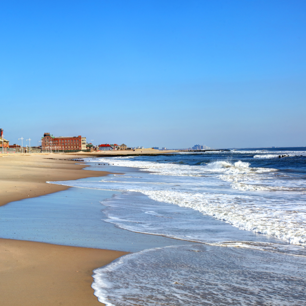 Waves reach the shore at Jacob Riis Park Beach in Queens, with wide sandy stretches, historic brick buildings, and a clear blue sky along the Atlantic coast.