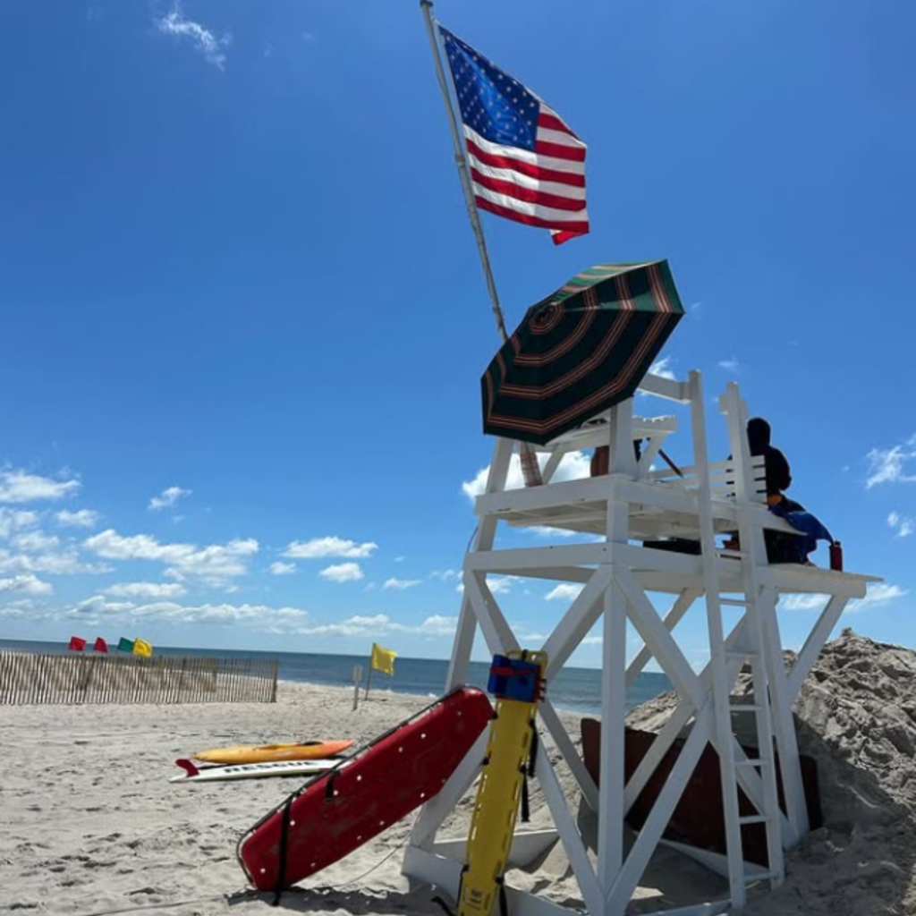 A lifeguard sits on a tall white stand at Jones Beach State Park in Wantagh, with rescue gear, an American flag, and colorful beach flags under a sunny sky.
