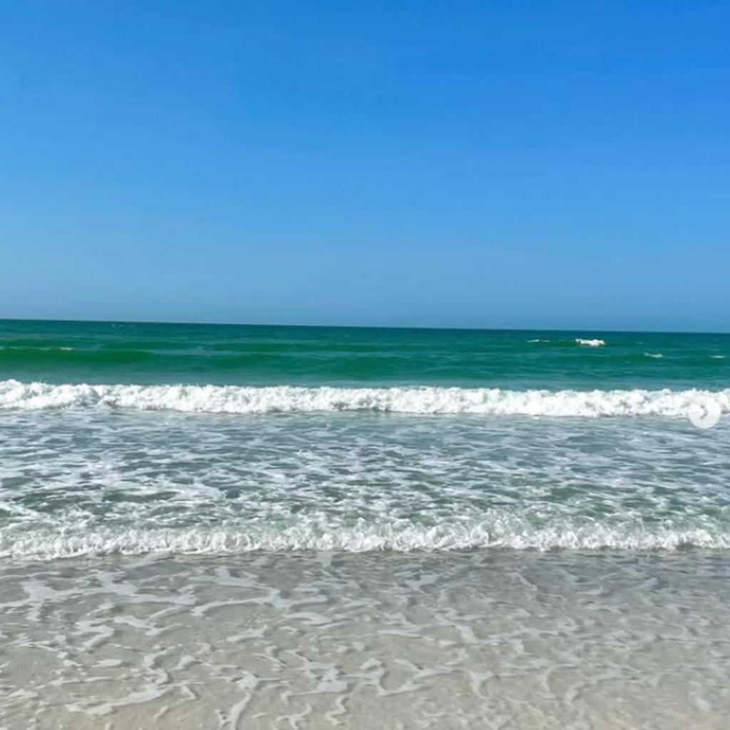 Clear waves roll onto the shore at Lido Beach in Lido Beach, New York, with emerald green water, gentle foam, and a bright blue summer sky above.