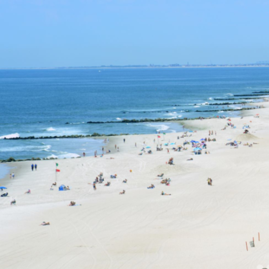 A wide view of Long Beach in Long Beach, New York, shows beachgoers spread across the soft sand with umbrellas, gentle waves, and rock jetties along the shore.