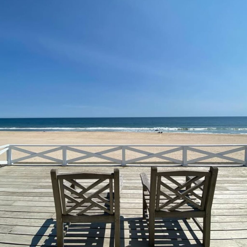 Two wooden chairs on a sunlit deck overlook a wide sandy beach and calm ocean waves under a clear blue sky, offering a peaceful coastal view.