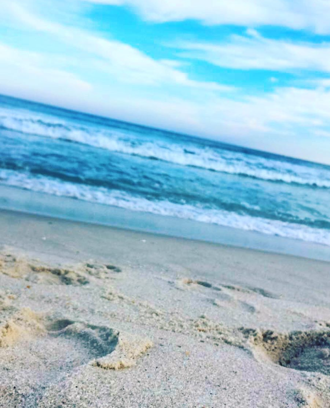 Close-up view of sandy footprints leading to gentle ocean waves under a partly cloudy blue sky at a peaceful beach with calm, rolling surf.








