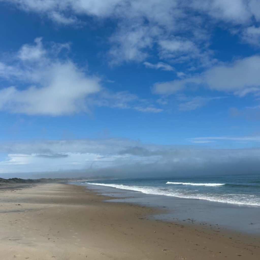 Waves gently meet the empty shoreline below Mohegan Bluffs on Block Island, RI, with soft sand, distant footprints, and a mix of blue sky and scattered clouds.