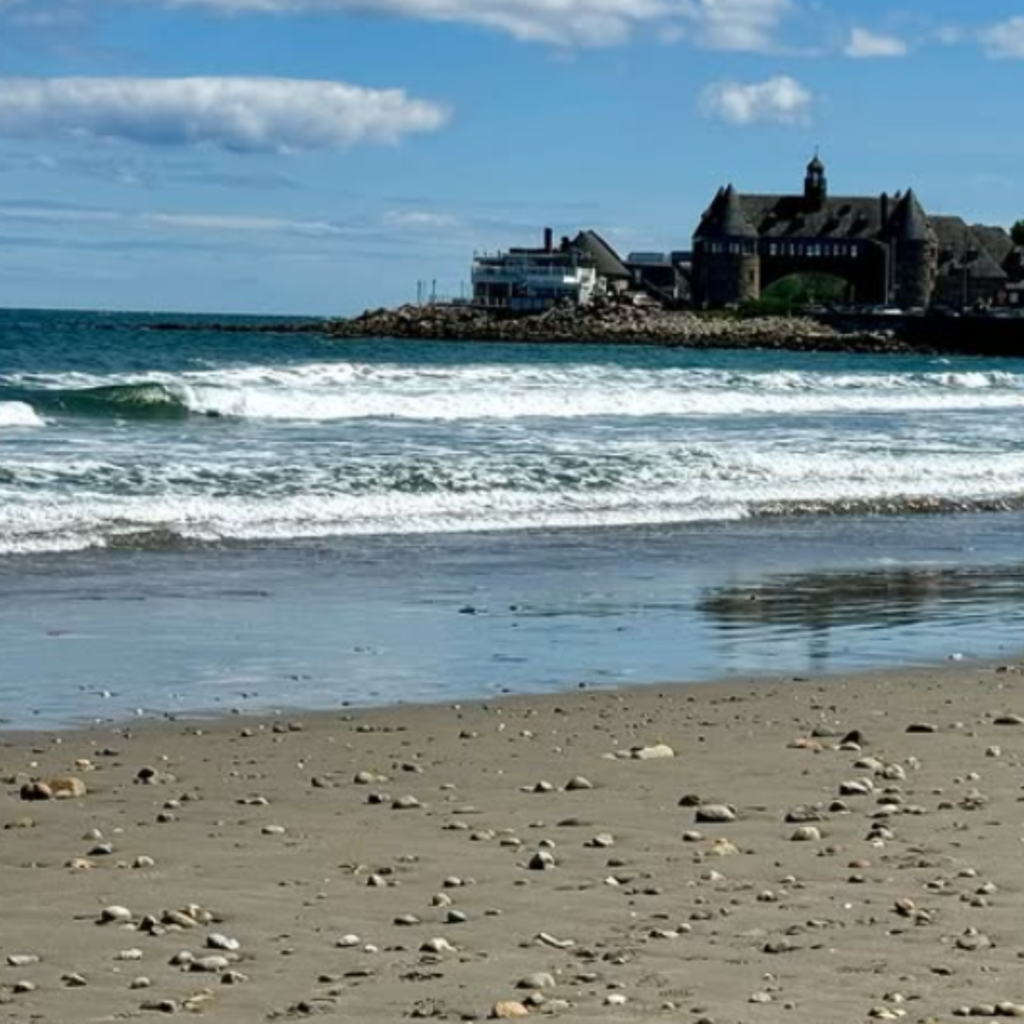 Waves roll onto the pebbled shore at Narragansett Town Beach in RI, with a historic stone building and rocky jetty visible in the distance under a sunny sky.