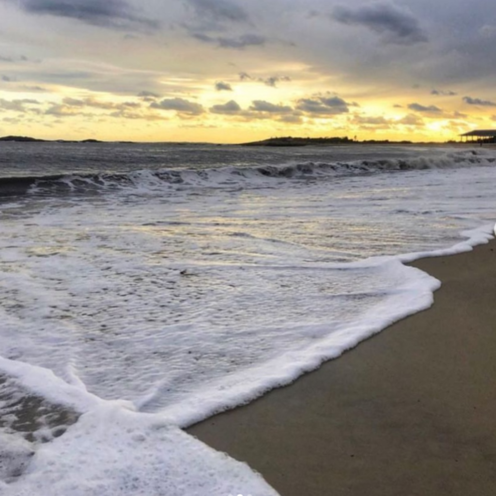 Waves roll onto the wet sand at Ocean Beach Park in New London, CT, as the sun sets behind distant clouds, casting a golden glow across the horizon.