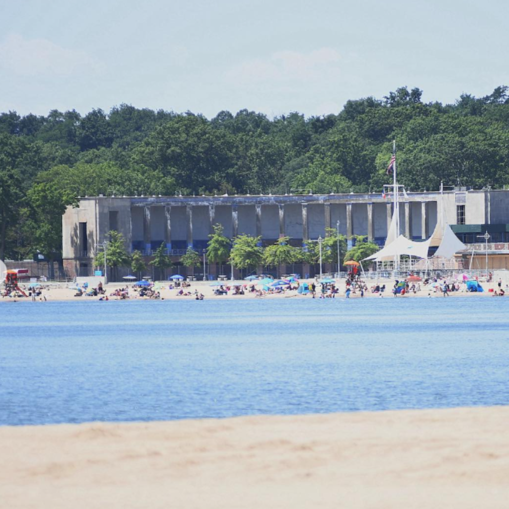Crowds relax along the sandy shore of Orchard Beach in the Bronx, with umbrellas, swimmers, and a large concrete pavilion surrounded by trees in the background.