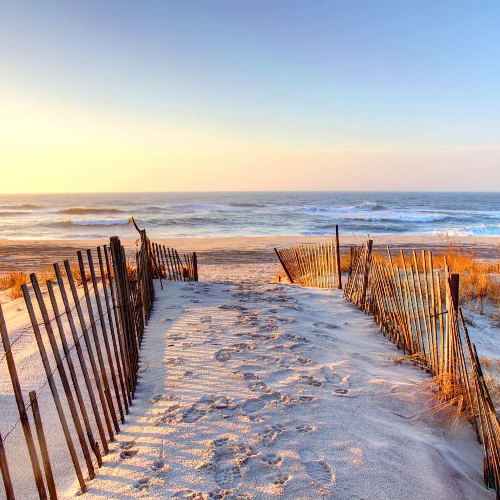 A sandy path leads to the shoreline at Ponquogue Beach in Hampton Bays, framed by wooden dune fences and golden sunrise light over calm ocean waves.