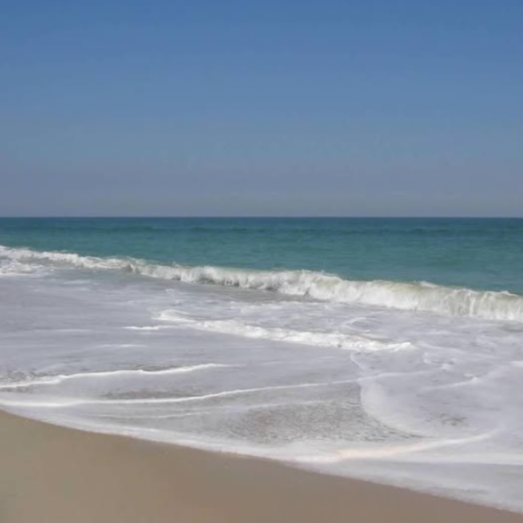 Gentle waves wash over the soft sand at Robert Moses State Park on Fire Island, with clear turquoise water and a sunny blue sky creating a peaceful view.