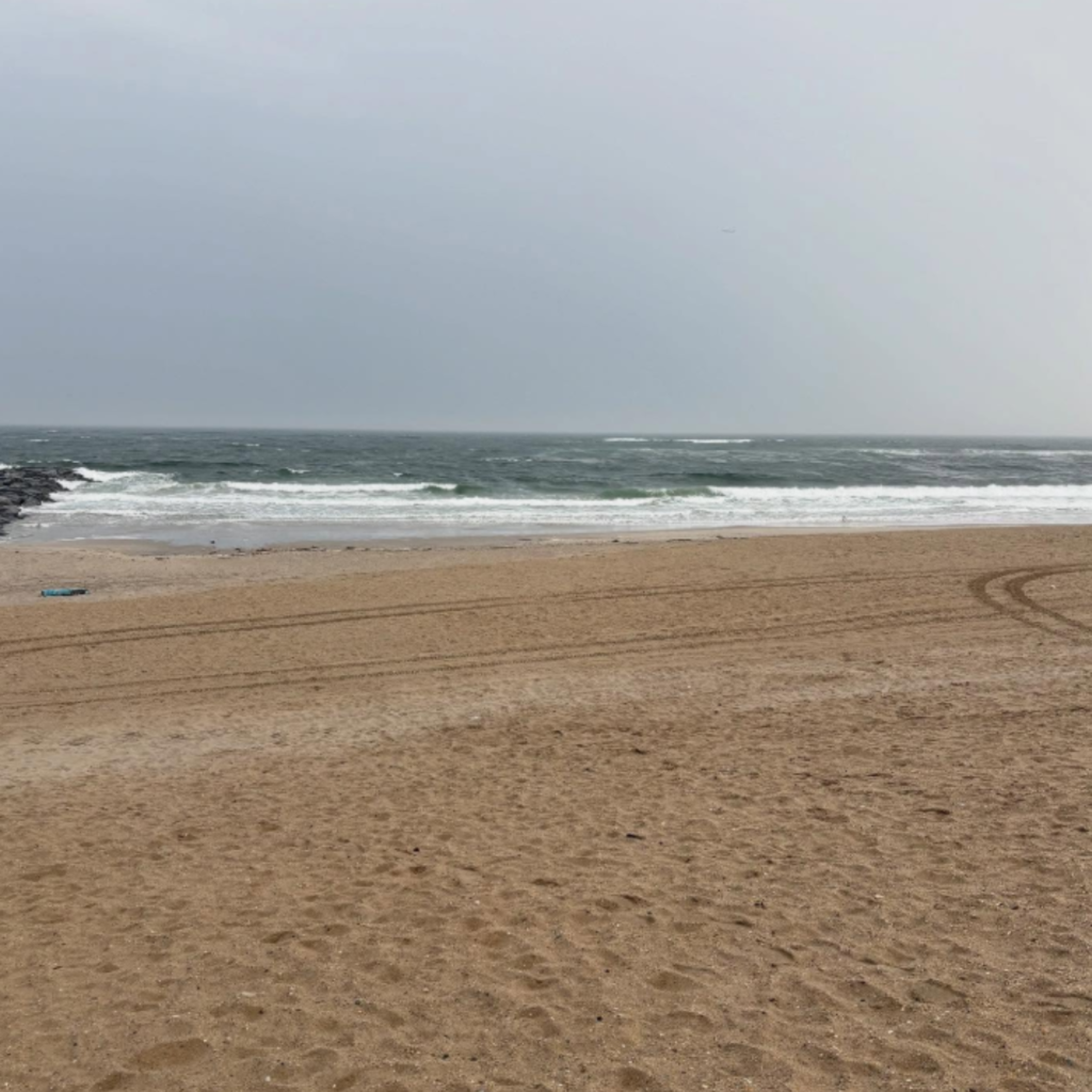 Overcast skies hang above Rockaway Beach in Queens, where waves crash onto the wet sand and tire tracks mark the wide, empty shoreline near a rock jetty.