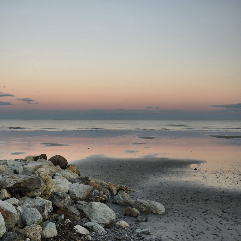 A peaceful view of Rye Town Park and Beach at low tide, with scattered rocks in the foreground and soft pink and blue sunset colors reflecting on the wet sand.