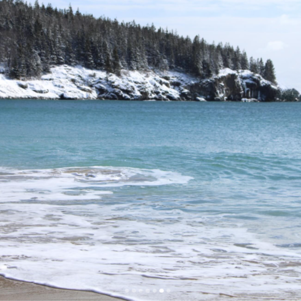 Waves gently wash onto the shore at Sand Beach in Acadia National Park, ME, with snow-covered cliffs and pine trees rising above the cold, clear blue water.