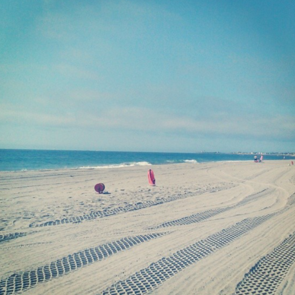 Fresh tire tracks curve across the soft sand at Scarborough Beach State Park in Narragansett, RI, with calm waves, lifeguard floats, and a hazy blue sky.