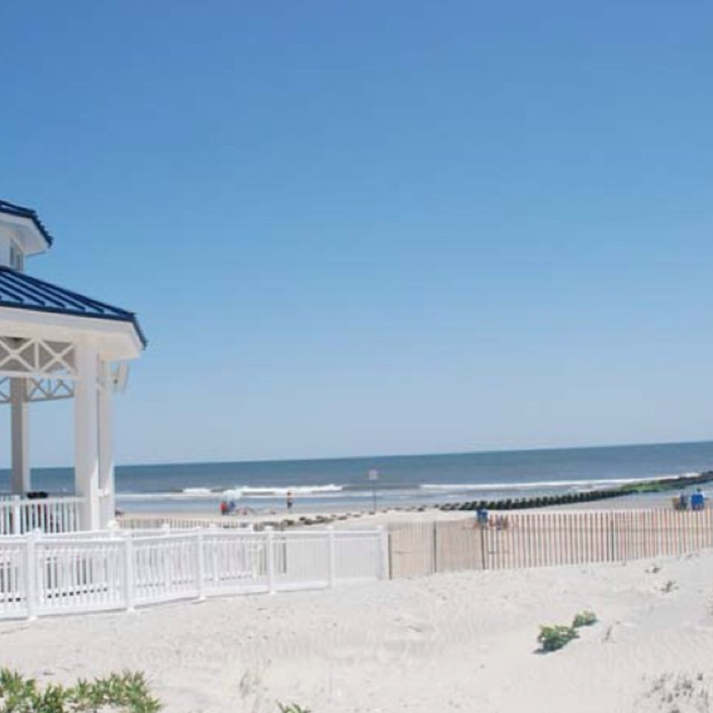White pavilion with a blue roof overlooks a peaceful sandy beach and calm ocean waves, under a clear blue sky in Sea Isle City, New Jersey.








