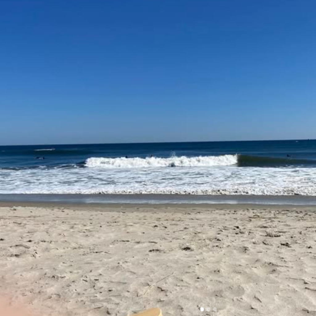 Wide sandy shore with gentle waves rolling in under a clear blue sky at Seabrook Beach, NH. A peaceful, sunny day with surfers in the distance.