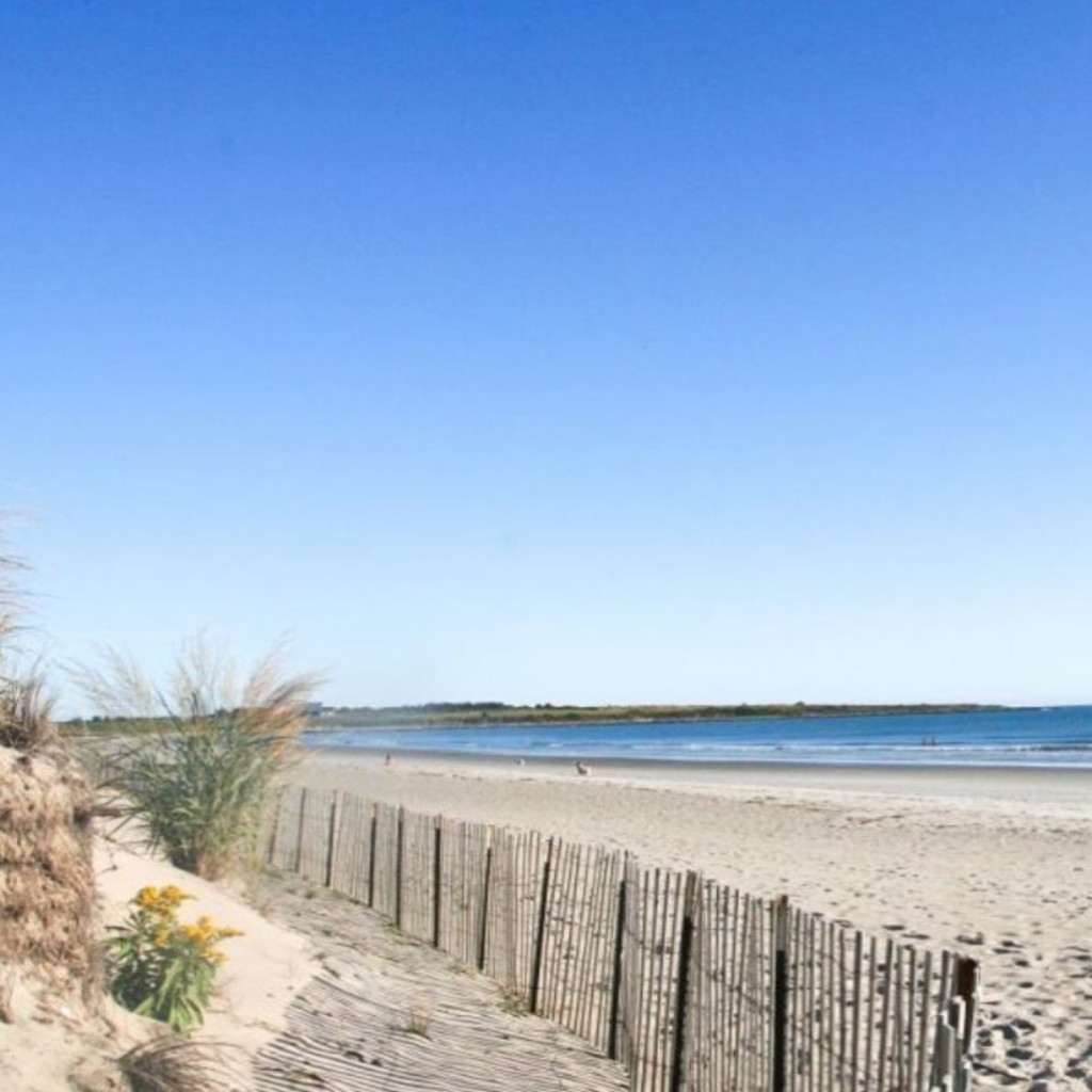 A sunny view of Second Beach in Middletown, RI, shows a sandy shoreline bordered by beach grass and wooden fencing, with calm waves and a clear blue sky.