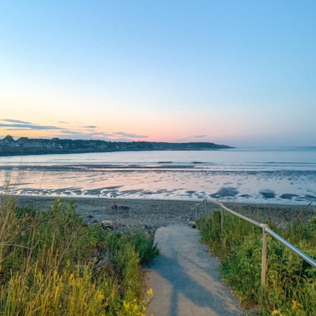 A sandy path lined with grass and railings leads to Short Sands Beach in York, ME, at sunset, with calm waves and soft pastel colors in the sky.
